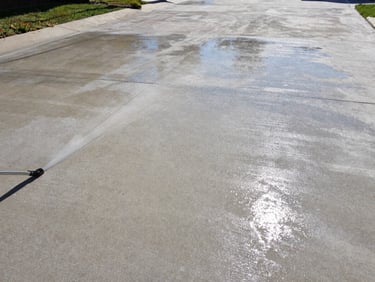 Wide environmental shot of the same concrete driveway after high-pressure cleaning, surface gleaming pale grey under midday sun, wet sheen reflecting light, completely free of algae and dirt, no people, full driveway width visible