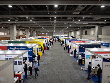 Wide environmental shot of a trade expo hall in India, rows of logistics and manufacturing supplier booths stretching into the distance under bright overhead lighting, documentary framing from a mid-height vantage point showing the scale of the floor
