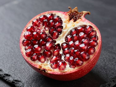 Studio macro photograph of a halved pomegranate on a dark slate surface, bright diffused overhead light, deep ruby arils densely packed and glistening, cross-section fully visible, no human subjects, no text