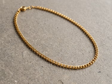 Medium shot of a finished 22-karat gold necklace laid flat on a matte warm-grey stone surface, north-facing indirect daylight revealing the precise chain construction and surface texture, no hands or props