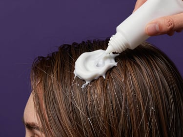 Close-up of a white treatment cream being smoothed through damaged, dry hair strands against a deep purple background, hands partially visible, studio fill light, no text