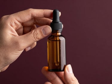 Amber glass dropper bottle of hair oil held between two fingers against a dark aubergine background, warm studio light catching the golden liquid, no text