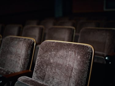 Dramatic low-key 35mm film still, empty cinematic theater seats in deep shadow, soft light catching the edge of a velvet chair, muted analog colors