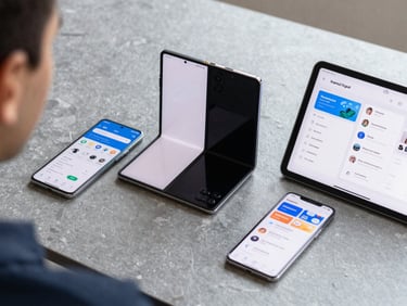 Over-the-shoulder environmental shot of three modern Android devices—a budget phone, a flagship foldable, and a tablet—lying on a slate grey desk, displaying synchronized responsive app layouts under bright neutral light.