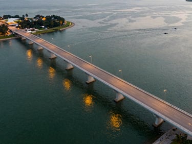 Aerial view of the Singapore-Johor causeway at dusk, golden lights reflecting on the water, clean architectural lines, warm evening light