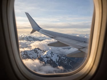 A clean airplane window view showing a silver wing cutting through golden hour clouds over snowcapped mountain peaks.