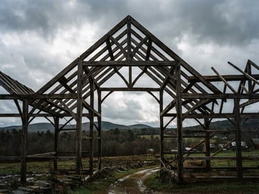 Wide environmental shot of a massive white oak timber frame silhouetted against a moody overcast Vermont sky, dramatic structural geometry, raw wood textures, 35mm film grain