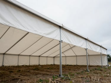 Wide environmental shot of a large white traditional marquee erected in an open field, heavy-duty PVC roof panels tensioned across steel frame poles, natural overcast daylight, no people, focus on scale and fabric tension