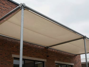 Wide shot of a bespoke architectural fabric canopy fixed to the rear elevation of a commercial building, fabric panels under tension between steel posts, overcast daylight, brick wall visible behind