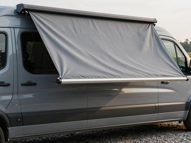 Side-on view of a van with a bespoke grey awning extended over a gravel pitch, awning fabric taut in afternoon light, vehicle bodywork and awning attachment rail in sharp focus