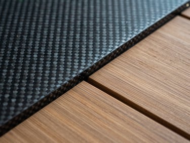 Close-up of raw marine materials including brushed teak decking meeting a carbon fiber weave panel under cool-toned technical lighting