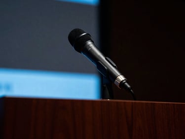 Tight, intense close-up of a corporate microphone on a dark wooden podium, shallow depth of field, cool blue screen-glow accents, moody charcoal tones.