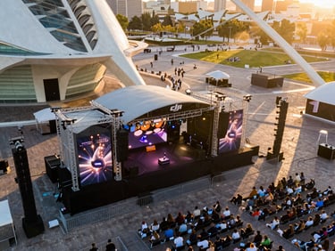 Aerial close-wide view of an outdoor brand event in Valencia, branded stage structure and audience grid visible from above, late afternoon golden light raking across the space, DJI Mini 5 Pro perspective, no faces identifiable