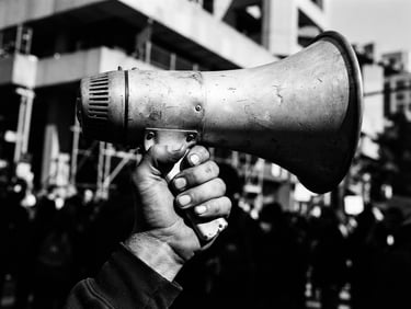 High-contrast black and white photograph, harsh direct sunlight, deep shadows, sharp focus, 35mm film grain, extreme close-up of a hand holding a vintage megaphone, urban architecture with strong lines in the blurred background, dynamic composition.