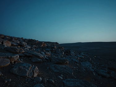 Poetic cinematic film still, wide shot of a rocky Palestinian landscape at twilight, soft neon blue horizon, minimalist framing, high contrast