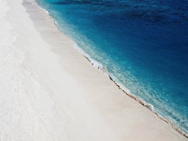High-angle minimalist shot of deep blue ocean water meeting white sand on a completely deserted tropical island shore.