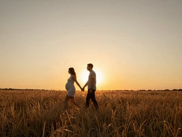 Wide cinematic shot of an expecting couple walking through a golden wild grass field at sunset, low horizon, expansive warm sky, natural unposed movement.
