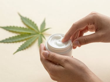 Hands gently applying a small amount of botanical cream from a jar onto skin, clean hemp leaf in the background, soft diffused morning daylight, shallow depth of field.