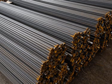 Wide overhead shot of a dense stack of TMT bars in a warehouse, corrugated ribs sharply visible under overhead work-lights, organized rows receding into the background, industrial floor visible at frame edge