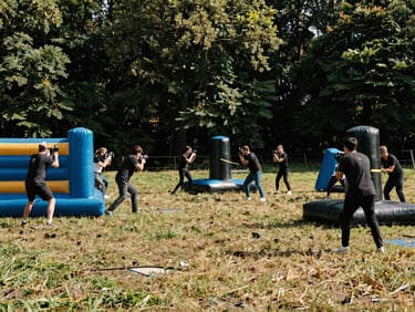 An outdoor laser game arena set up on a public grassy square with inflatable obstacles, players in action under bright daylight.