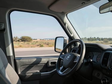 Wide shot of a vehicle interior from the passenger side, steering wheel and open highway ahead under direct midday sun, dashboard instruments visible, no driver face shown