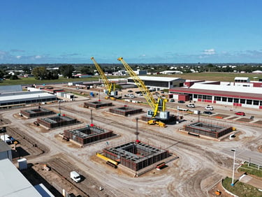 Vue aérienne d'un site de génie civil avec des fondations en béton et des grues, sous un ciel bleu éclatant, style industriel et précis.