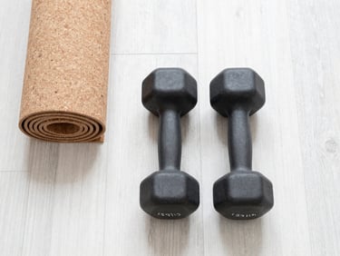 Top-down flat lay of a matte black dumbbell and a cork yoga mat on a bright white oak floor, soft morning daylight, airy neutral tones, 35mm