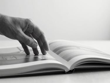 A close-up of a hand gently turning the page of a high-quality art book, soft natural diffused light, desaturated black and white, sharp focus, 35mm architectural lens.
