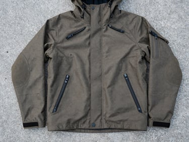 Flat-lay composition of a rugged tactical jacket laid out on cold concrete, showing heavy zippers and reinforced elbows, desaturated tones