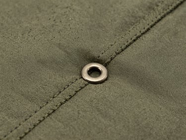 Extreme close-up macro shot of double-stitched olive-drab seams on heavy nylon fabric, metallic grommet visible, hard overcast lighting showing grit and texture