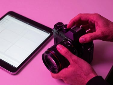 Vibrant close-up of a designer's hands adjusting a 35mm camera lens next to a tablet showing clean UI wireframes, high-contrast pink neon lighting, cinematic shadows.