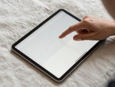 Close overhead shot of a person's hands scrolling a simple website on a tablet resting on a cream linen surface, natural overcast daylight, one finger hovering over a selection