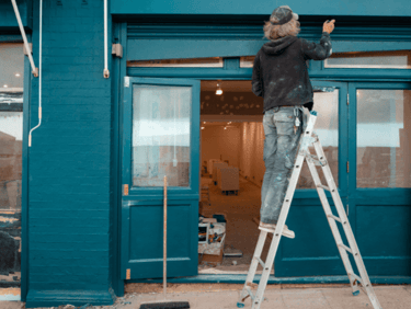 A professional painter on a ladder painting a blue storefront during a shop renovation.