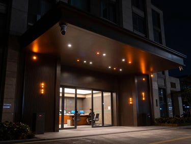 Dramatic low-key wide shot of a modern condominium entrance at night, sharp focus on high-tech surveillance cameras with safety orange status LEDs, security command center feel.