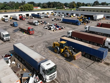 Overhead view of a scrapyard facility showing multiple trucks and a loader in operation under open sky, organized material zones visible, scale of the working yard apparent from the elevated angle