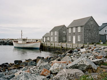 Weathered shingle architecture along a quiet, rocky coastline under soft overcast daylight, with a historic wooden fishing boat in a calm harbor.