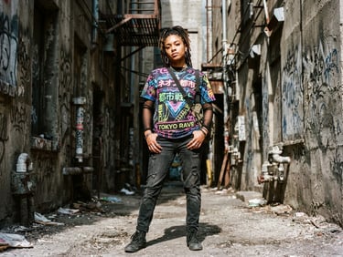 Gritty urban fashion photography, harsh direct sunlight, a young creative wearing a vibrant sublimated streetwear t-shirt with neon graphics, standing in a concrete industrial alley, raw texture, 35mm film grain
