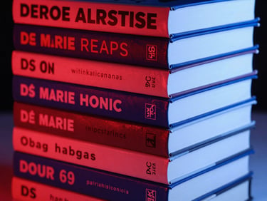 Graphic close-up of a stack of independent paperback books with bold typography on the spines, high-contrast studio lighting, red and blue gel accents.