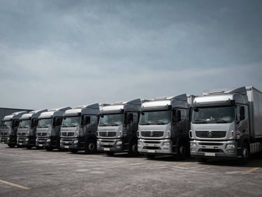 A row of newly manufactured commercial transport trucks parked at a logistics depot, high-contrast industrial lighting, desaturated steel tones, wide-angle framing.