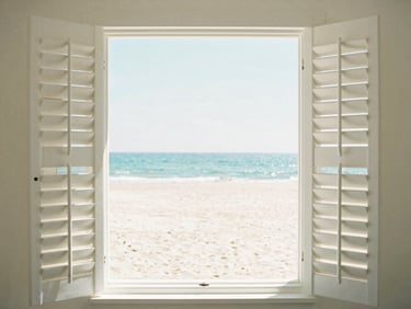 Crisp white plantation shutters fitted on a large window overlooking a bright sandy beach scene.