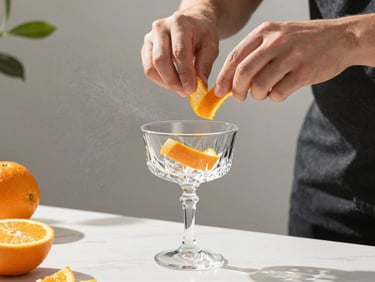 Hands actively expressing a fresh orange peel over a crystal coupe glass, fine mist visible in soft sun-dappled daylight, creative mixology setting