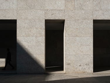 Cinematic architectural detail in Madrid, harsh midday shadows cast on minimalist stone facade, high contrast, raw textures of granite, human figure walking past in the distance, small in frame.