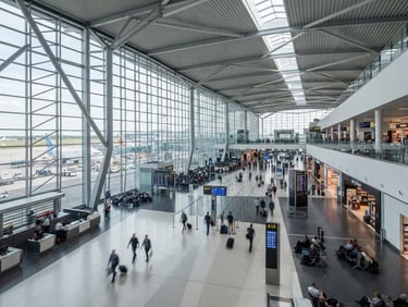 Fotografía arquitectónica de un aeropuerto moderno con luz natural brillante, líneas geométricas limpias, tonos fríos desaturados, mostrando escala y flujo humano ordenado.