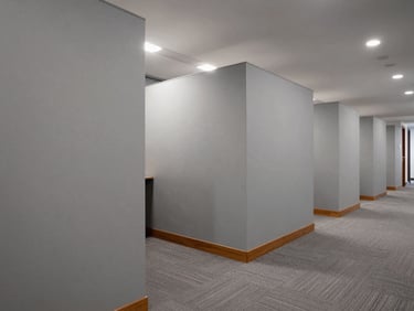 Wide-angle view of a corporate office corridor with solid opaque partition walls creating defined private office zones, neutral gray surface finish, warm wood skirting detail, soft overcast office lighting, clean and functional framing