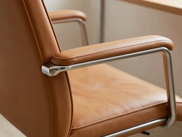Close-up of a director-class office chair armrest and side panel, warm tan leather upholstery texture with fine grain visible, polished aluminum trim catching soft window light, neutral background, material detail shot