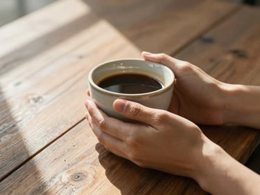Hands holding a warm ceramic coffee cup on a rustic wooden table, bright morning light spilling across the surface.
