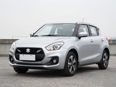 Front-three-quarter angle of a silver Suzuki compact car parked on a neutral outdoor asphalt surface under soft overcast daylight, sharp focus on the clean grill.
