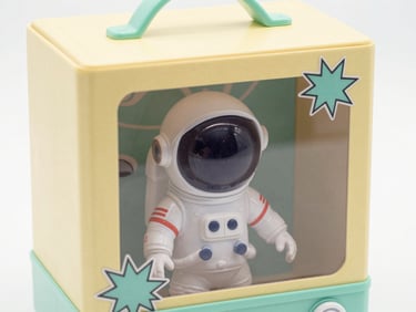 Close-up detail of a fake retro toy box design showing a tiny astronaut action figure in a bubble helmet, pastel yellow and mint packaging with comic-style starburst graphics, clean flat studio lighting