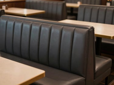 Empty restaurant booth with charcoal vinyl banquette seating, clean stitching visible along the back panel, warm interior lighting, tables set but no diners, no people