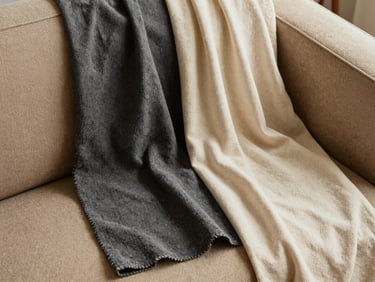 Overhead flat lay of fabric swatches in warm sand, charcoal and cream tones draped over a sofa armrest, texture and weave clearly visible under north-facing daylight, no people